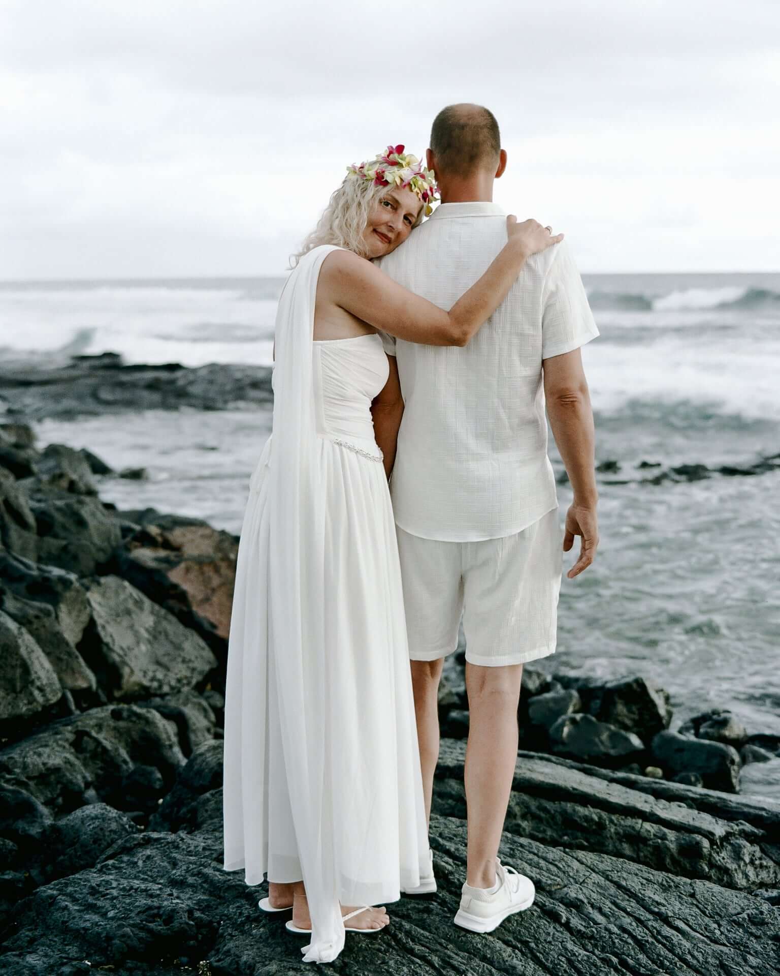 Couple embracing on lava rocks facing the ocean, the bride in a white dress with a flower crown