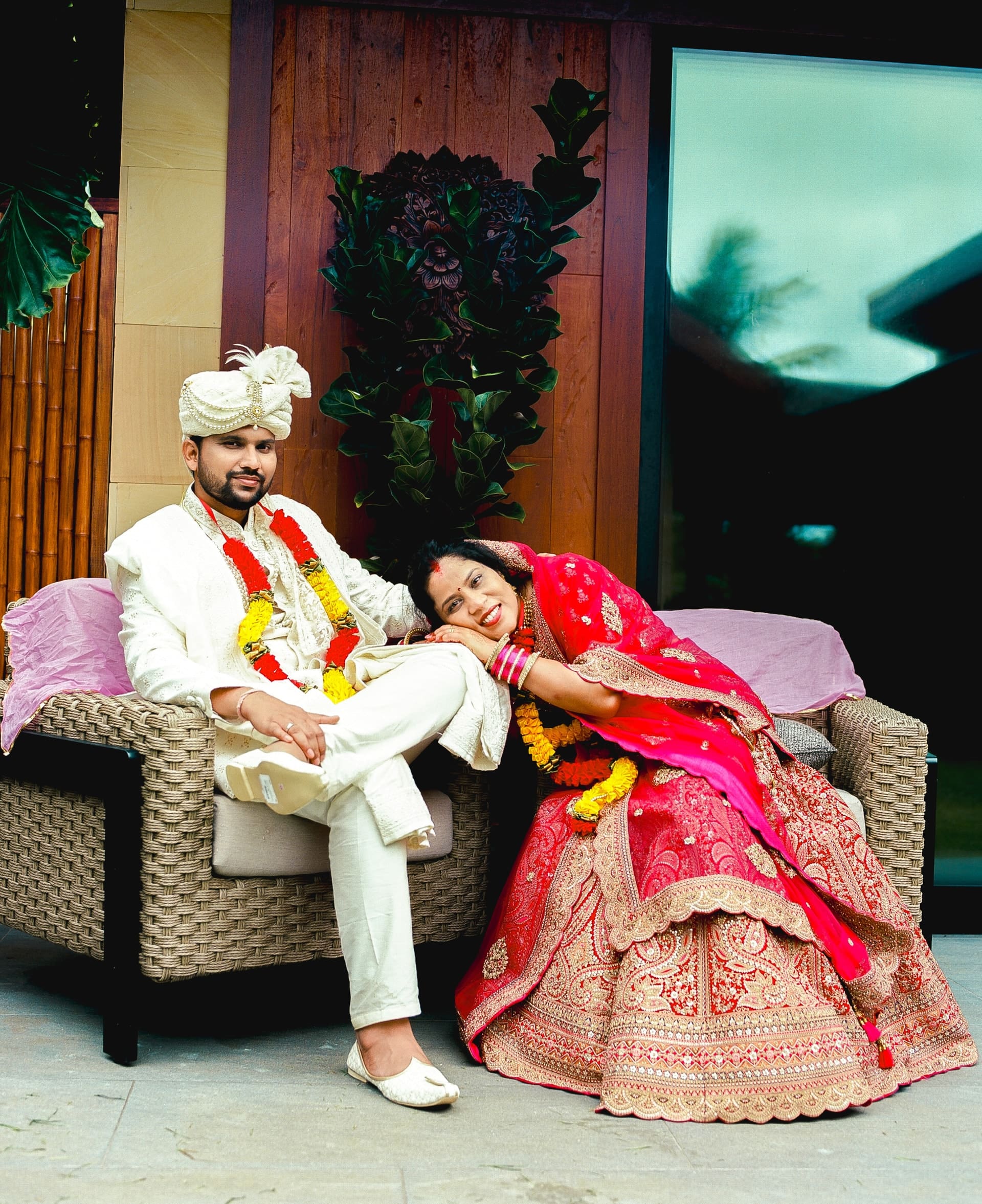 Groom in a white turban and bride in a pink lehenga sitting together on a rattan sofa