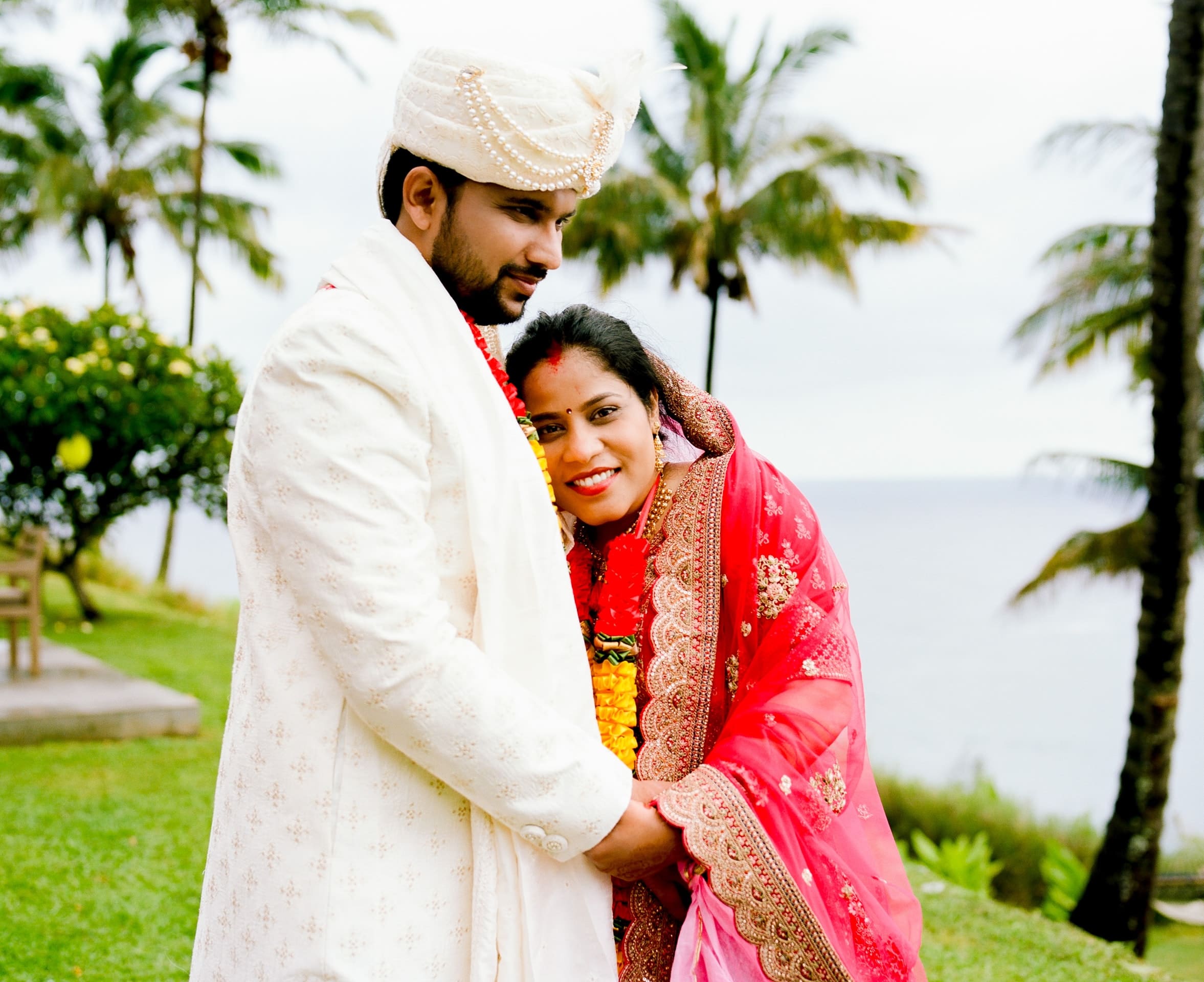 Groom in a white turban and bride in a pink sari smiling together by palm trees