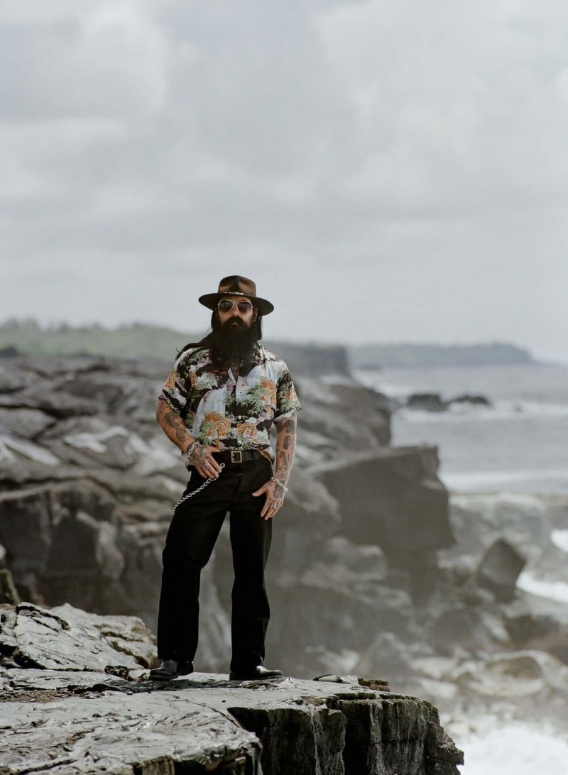 Bearded man in a hat standing on coastal rocks above the ocean