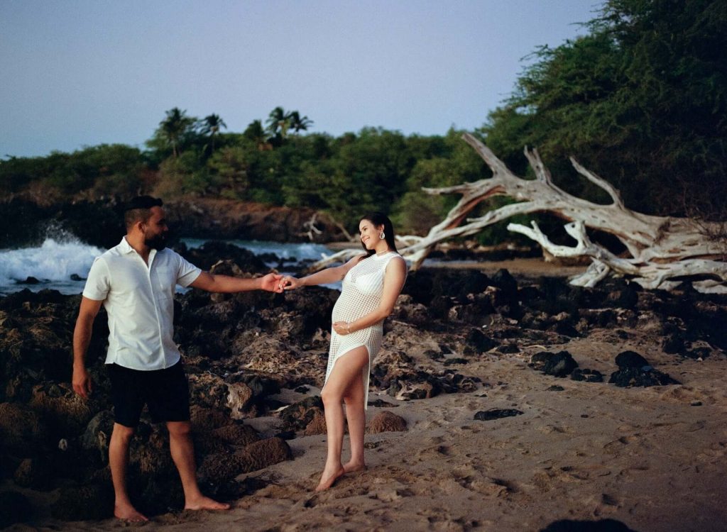 Couple holding hands between driftwood trees on the shoreline, maternity photographer in Kona