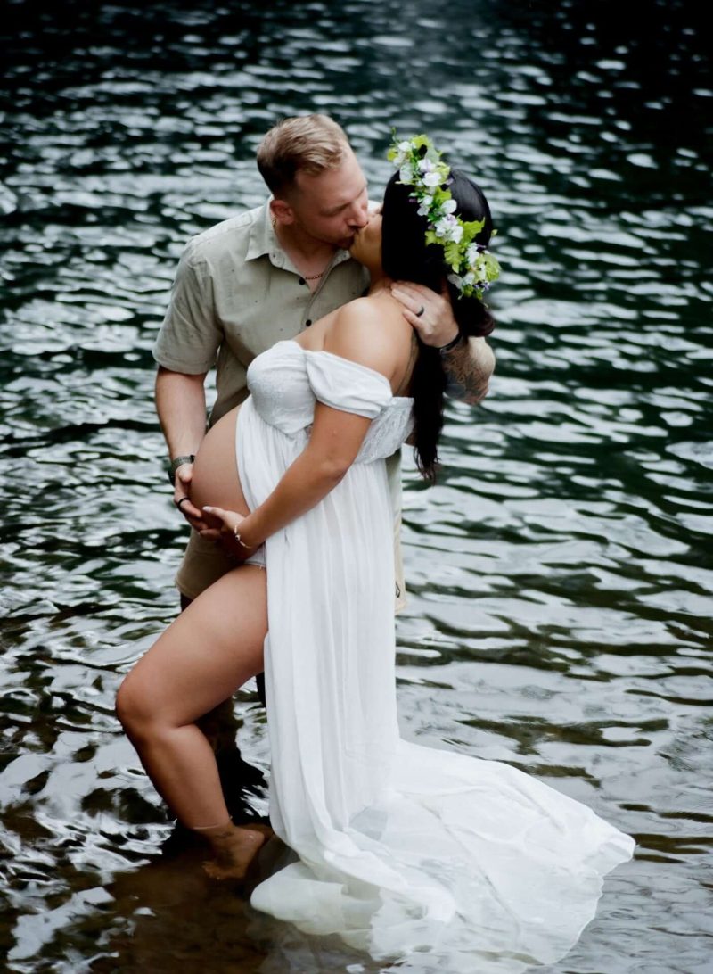 Couple embracing in a stream, the woman in a white maternity gown and flower crown