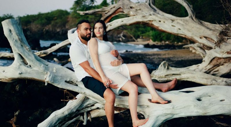 Expecting couple sitting on sun-bleached driftwood by the ocean