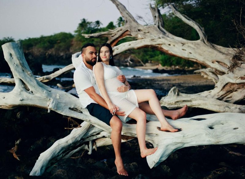Expecting couple sitting on sun-bleached driftwood by the ocean